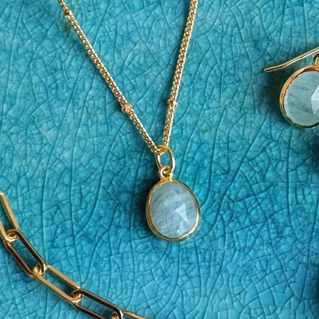 Niche Jewellery Sarah Alexander aquamarine gemstone pendant on a gold bobble chain against a blue textured background