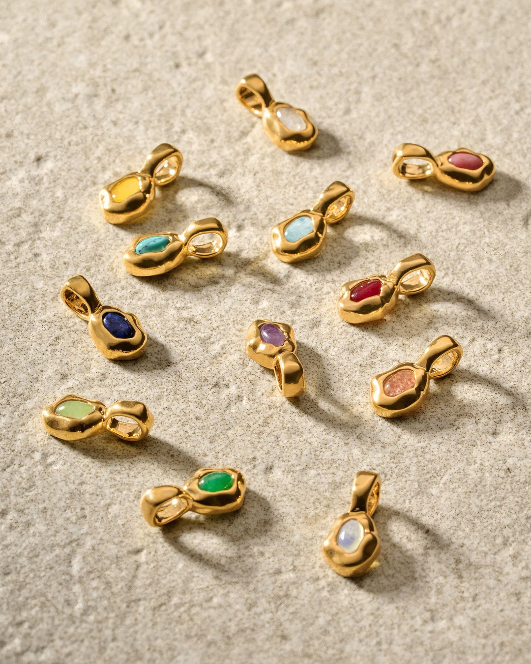 Niche Jewellery Katie Loxton gold birthstone pendants featuring various colourful gemstones on a textured surface