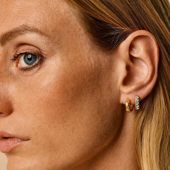Niche Jewellery Kali Charlotte's Web huggie hoops in 24ct gold plated brass with a faceted blue topaz stone worn on a woman's ear.