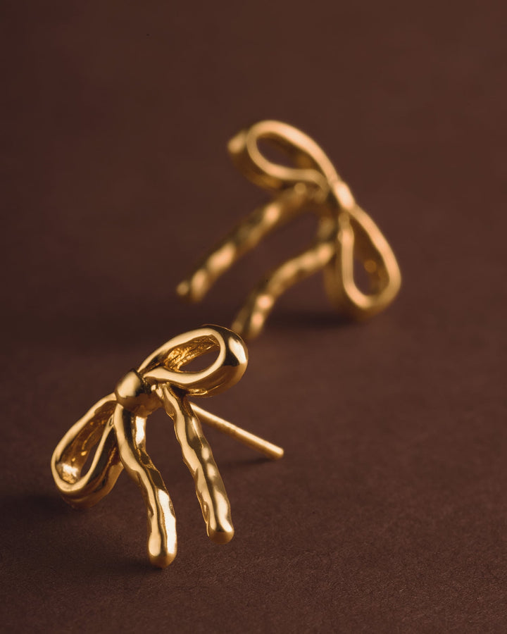 Niche Jewellery Katie Loxton Novelle waterproof gold-tone bow stud earrings on a dark brown background.