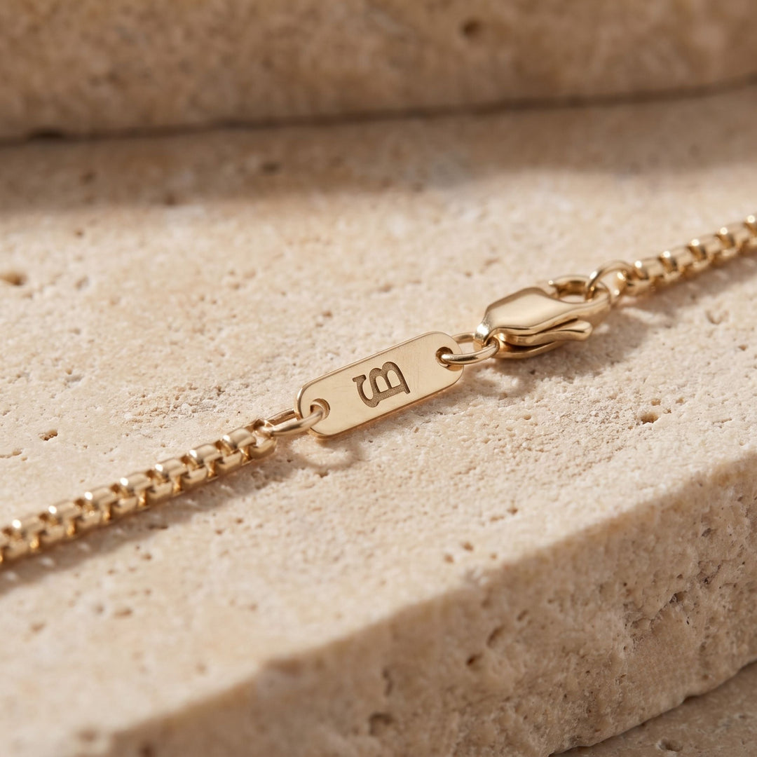 Niche Jewellery thin gold box chain necklace with branded logo tag and lobster clasp on a textured stone surface