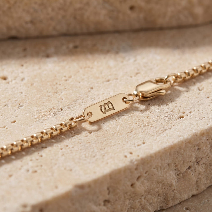 Niche Jewellery thin gold box chain necklace with branded logo tag and lobster clasp on a textured stone surface