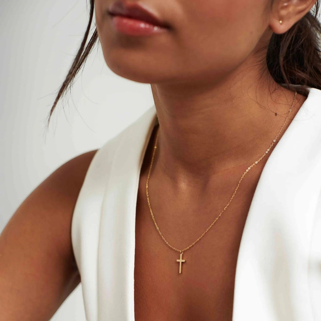 Niche Jewellery D.Louise gold cross pendant necklace on a fine chain worn by a woman in a white top.