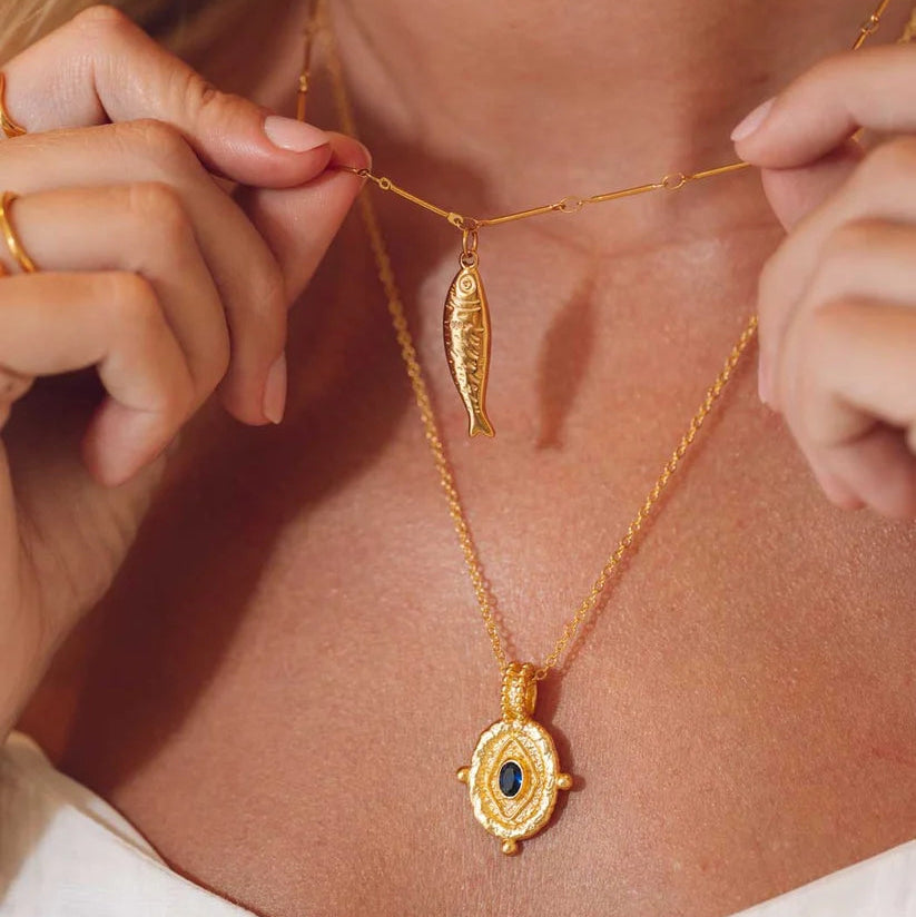 Niche Jewellery gold Selkie fish pendant necklace layered with a textured evil eye charm on a woman wearing white