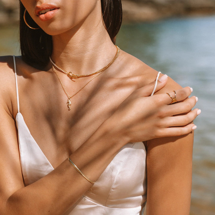 Niche Jewellery gold flat snake chain bracelet worn on a woman's wrist with a white silk dress by the water