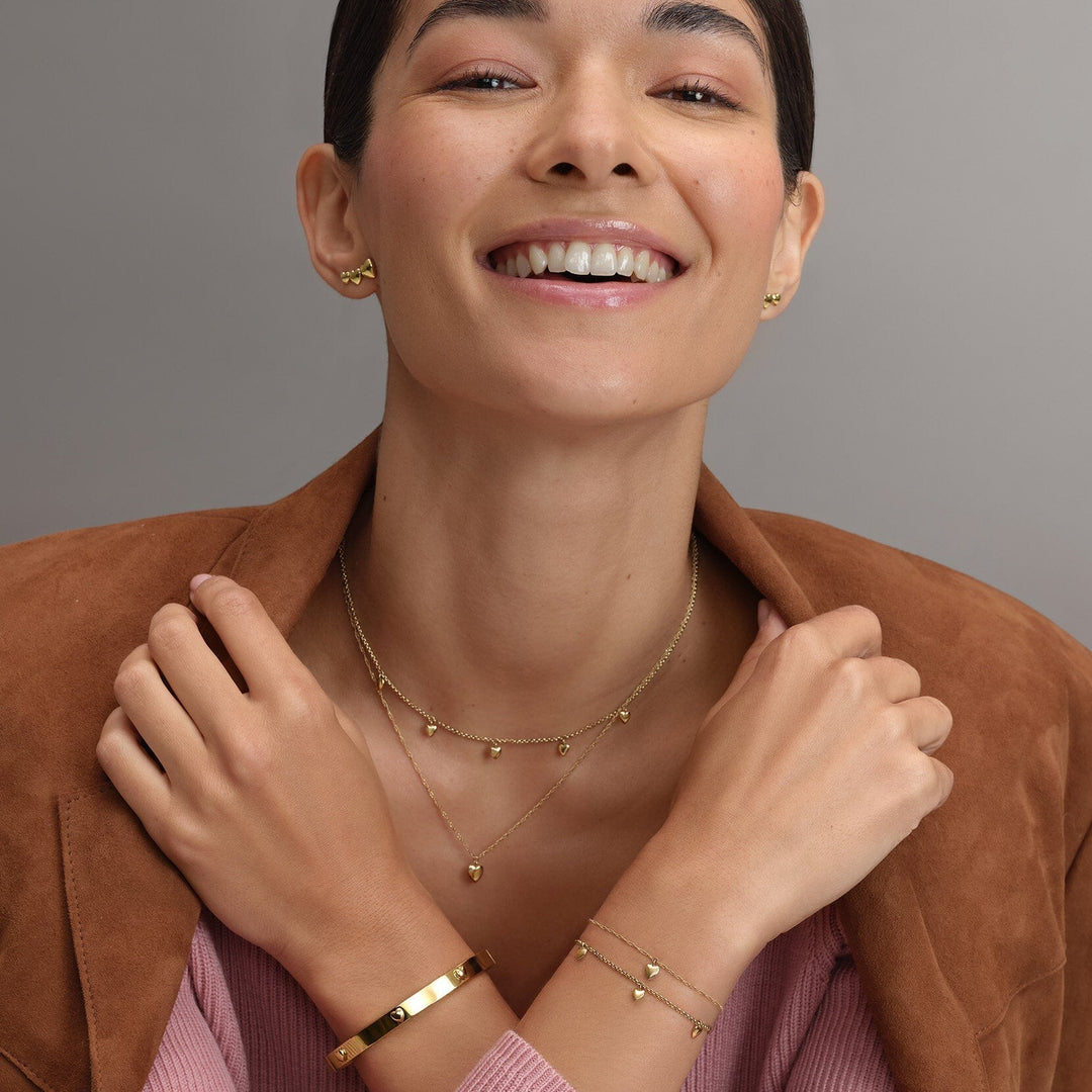 Niche Jewellery Olivia Burton gold plated bangle featuring a mini heart charm worn by a smiling woman in a brown blazer