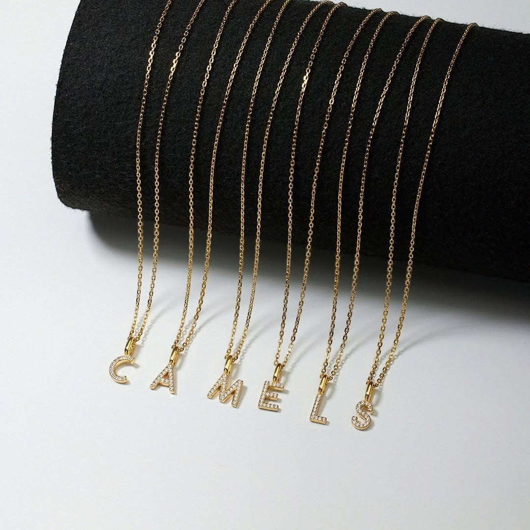 Niche Jewellery Ania Haie gold initial letter necklaces with pavé crystals on delicate chains displayed on a black stand.