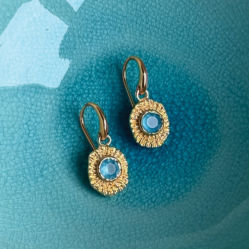 Niche Jewellery gold talisman drop earrings featuring a central blue gemstone on a textured sunburst setting.