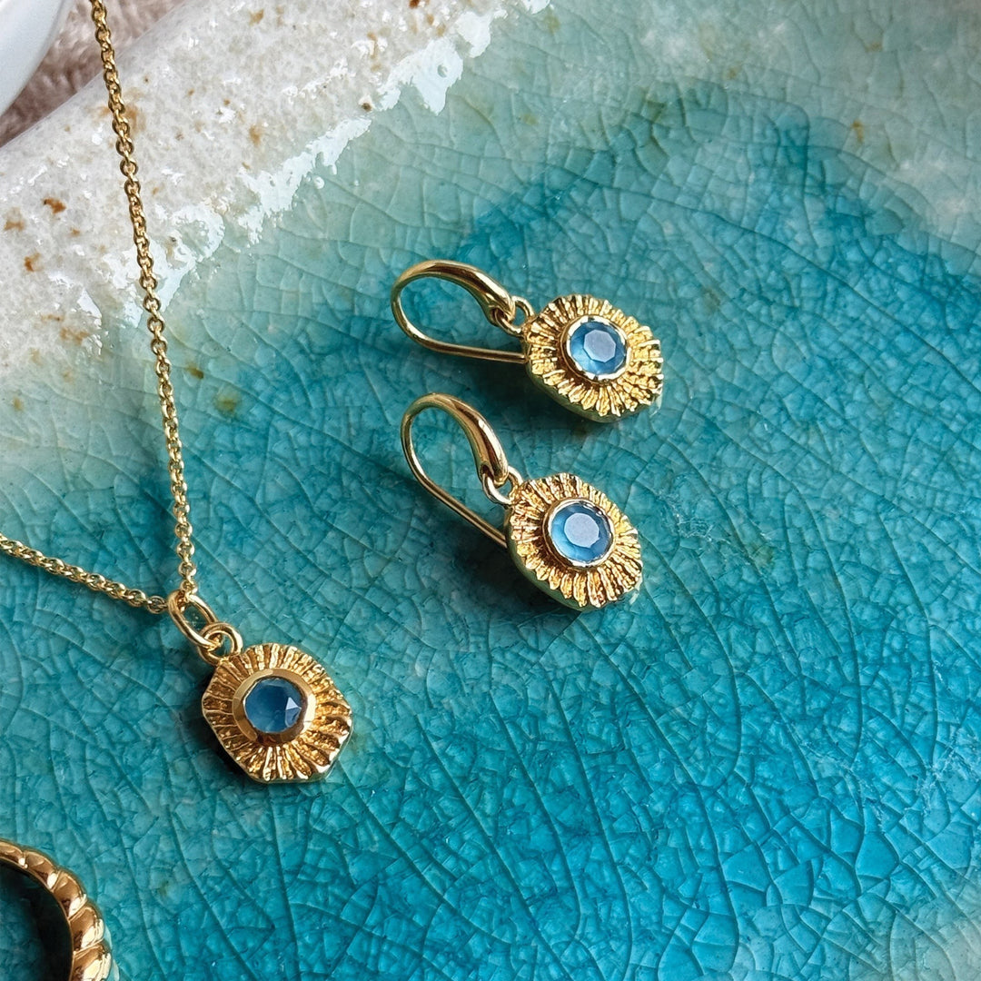 Niche Jewellery gold talisman drop earrings with blue gemstones on a textured turquoise ceramic dish