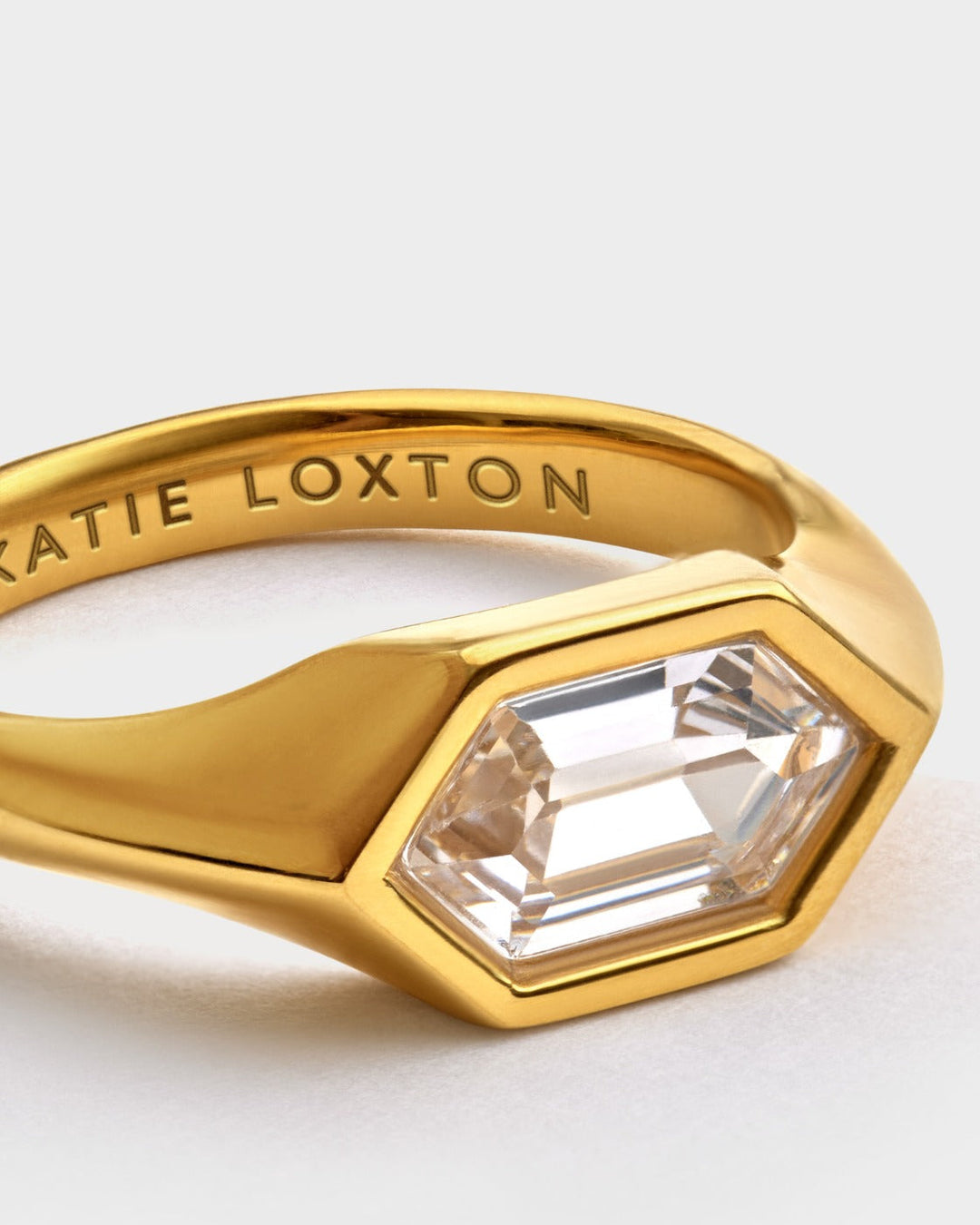Niche Jewellery Katie Loxton gold-tone waterproof statement ring with a hexagonal clear stone and engraved branding.