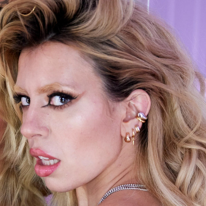 Niche Jewellery Scream Pretty mixed metal huggie earrings worn by a woman with blonde hair against a purple background.