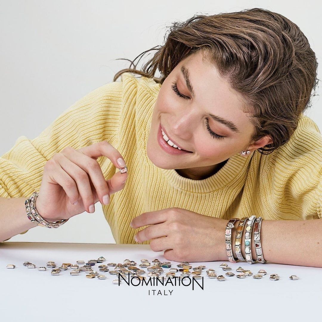 Niche Jewellery Nomination gold and light blue enamel forget-me-not charm held by a woman wearing a yellow sweater and bracelets.